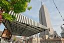 A black-and-white striped canopy covers a rooftop bar with people sitting beneath it. In the background, the tall Empire State Building rises against a blue sky in New York City. Tree branches frame the left side of the image.