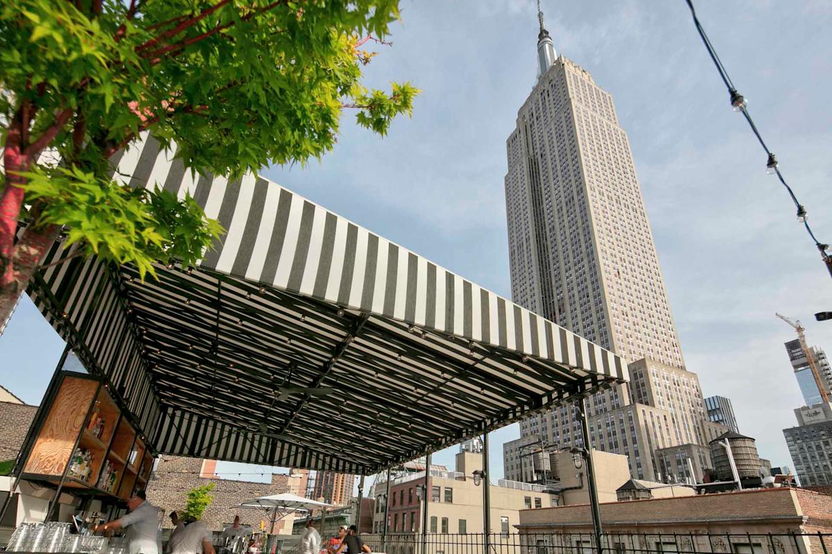 A black-and-white striped canopy covers a rooftop bar with people sitting beneath it. In the background, the tall Empire State Building rises against a blue sky in New York City. Tree branches frame the left side of the image.
