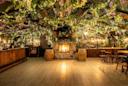 A cozy bar with wooden furniture and a stone fireplace, decorated with hanging greenery, flowers, and string lights creating a warm, inviting atmosphere. Two barrels stand near the fireplace, enhancing the rustic charm.