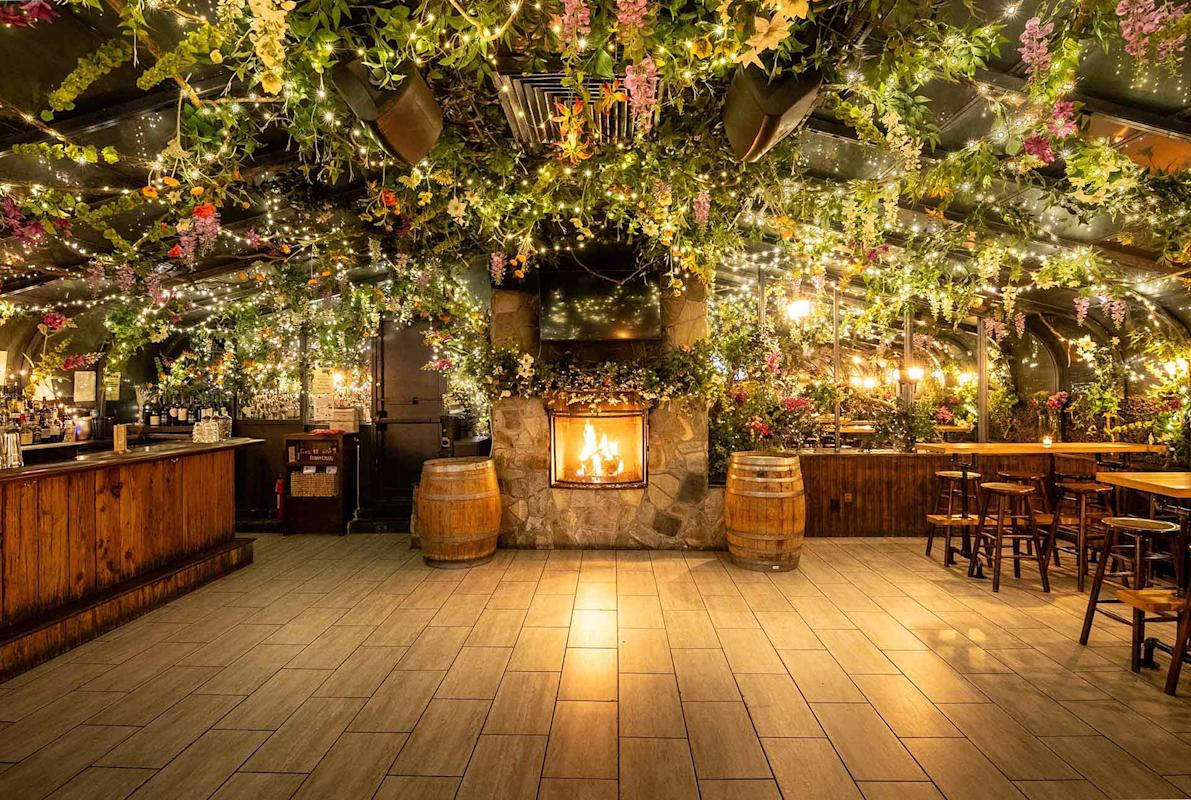 A cozy bar with wooden furniture and a stone fireplace, decorated with hanging greenery, flowers, and string lights creating a warm, inviting atmosphere. Two barrels stand near the fireplace, enhancing the rustic charm.