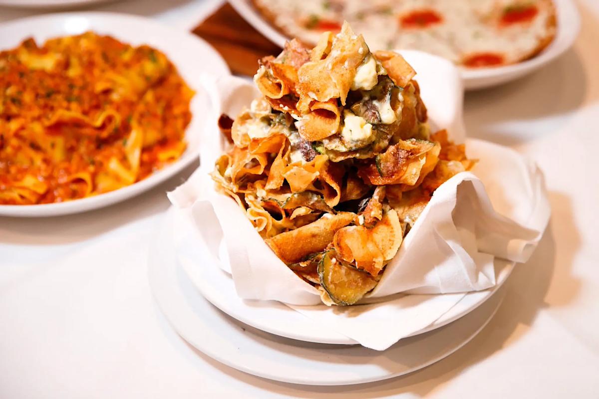 A plate of crispy fried vegetable chips with cheese is served on a white napkin, with dishes of pasta and pizza blurred in the background.