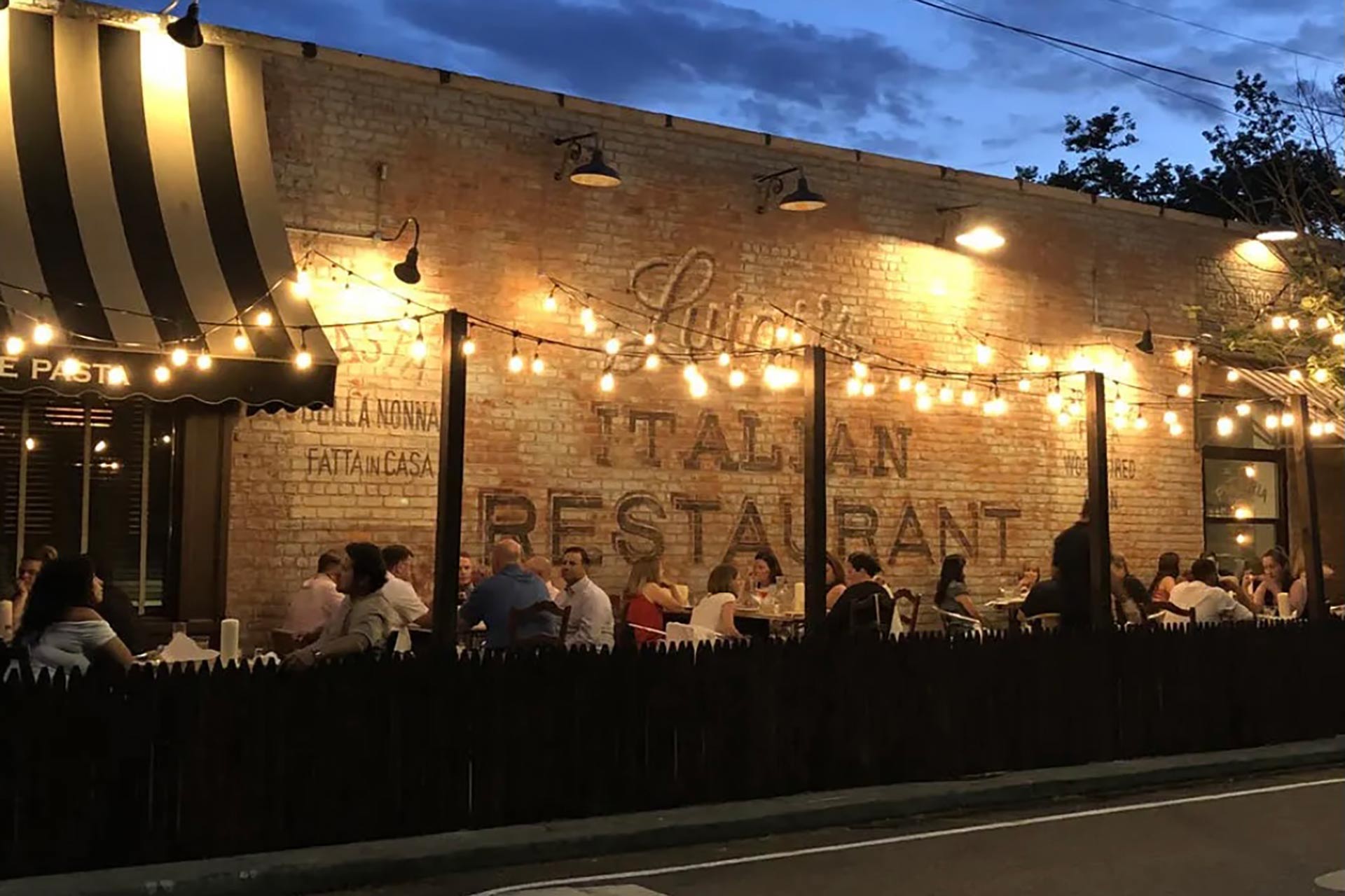 Outdoor dining area of an Italian restaurant at dusk, with string lights overhead, groups of people sitting at tables, and a brick wall with faded signage in the background.