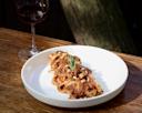 A white plate of pasta topped with a rich meat sauce, pine nuts, and a sprig of rosemary sits on a wooden table next to a glass of red wine.