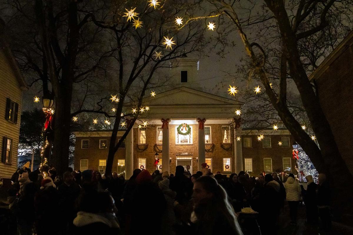 A crowd gathers at night in front of a large, decorated building with columns, illuminated by hanging star-shaped lights and festive wreaths, creating a warm holiday atmosphere.