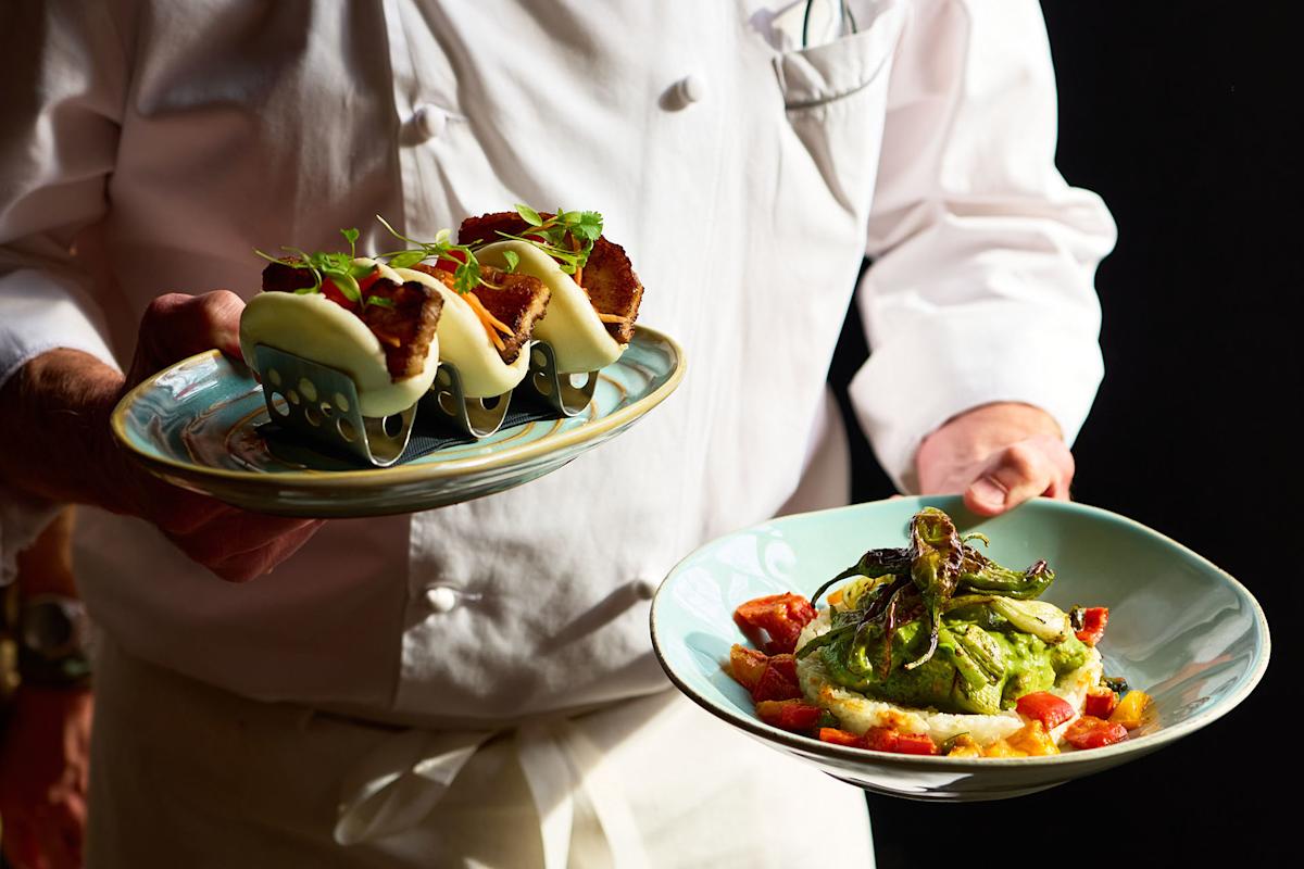 A chef in white uniform holds two plates: one with three tacos topped with cheese, the other with mashed potatoes, green sauce, grilled vegetables, and diced tomatoes.