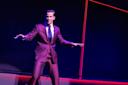 A man in a maroon suit and tie stands on stage with bent knees and arms extended, as if dancing or performing. The background is dark with bright red diagonal lines.