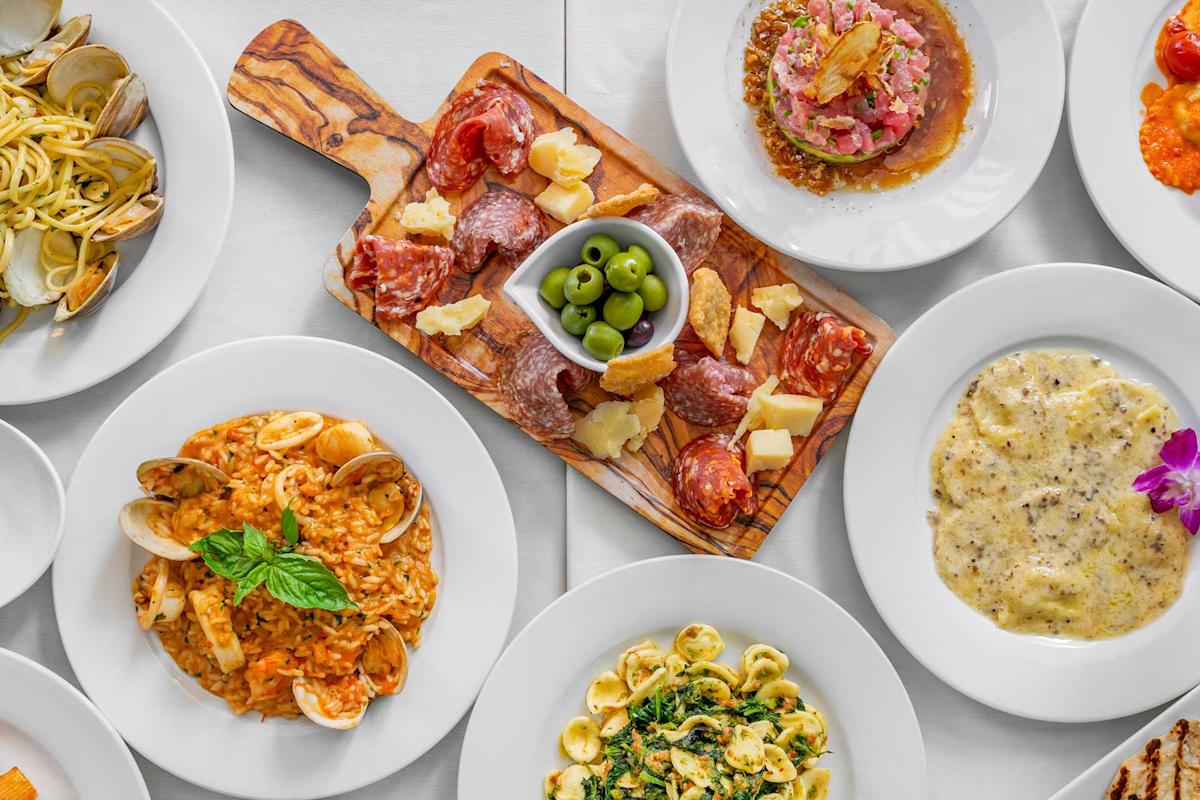 A table set with various Italian dishes, including pasta with clams, risotto, ravioli, orecchiette, a charcuterie board with cheese and cured meats, and a plate of marinated fish.
