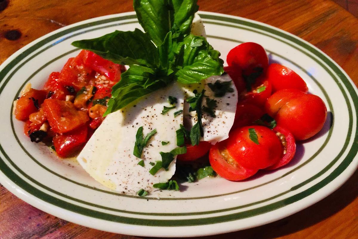 A plate of caprese salad with sliced mozzarella, fresh tomatoes, basil leaves, chopped herbs, and ground black pepper, served on a green and white striped dish.