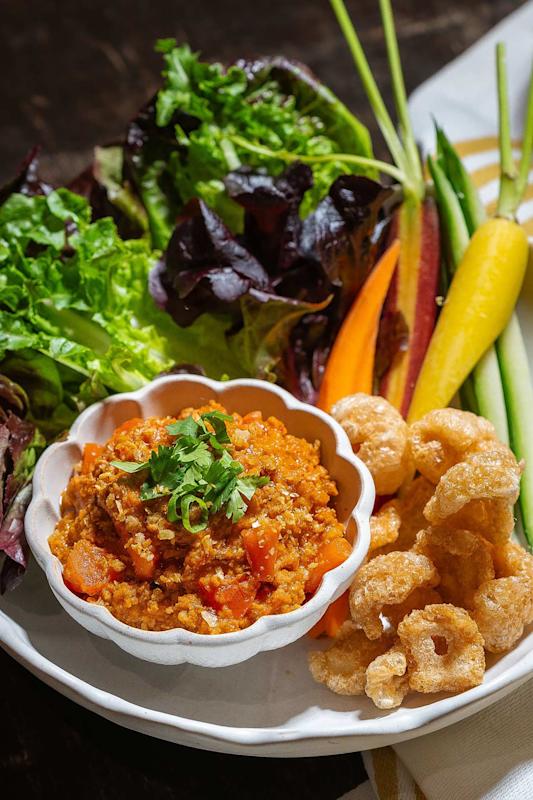 A plate with a bowl of spicy ground meat dip, fresh greens, colorful vegetable sticks, and crispy pork rinds, garnished with chopped cilantro.