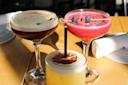Three cocktails on a wooden table: one creamy with floating coffee beans, another pink with blueberries on a skewer, and the third yellow with a dried citrus slice and a black straw. White napkins are in the background.