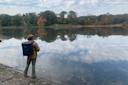 A person in outdoor clothing fishes by a calm lake, surrounded by autumn trees. The sky is reflected in the water, and the scene is peaceful and serene. The person carries a backpack and stands on a rocky shoreline.
