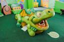 A large, friendly green dragon sculpture with wings and a wide open mouth sits on a green carpet in a colorful indoor children's play area. A speech bubble sign reads, "Hi! I'm Archie. Please feed me letters!.