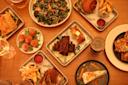 A variety of dishes on a wooden table, including fries, grilled meat, a salad, a burger, and dessert. Some plates have dipping sauces, and a drink is placed at the edge. The arrangement is casual and inviting.