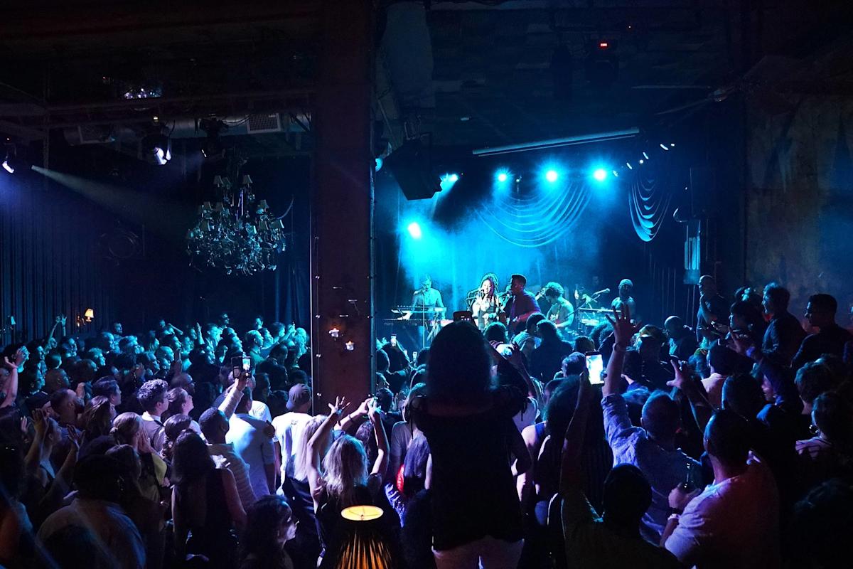 A crowded, lively concert scene with a band performing on stage under blue lights. The audience is standing closely together, many with hands raised, enjoying the music in a dimly lit indoor venue.