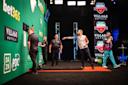 A professional darts match in progress, with one player throwing a dart, others watching, and a referee observing, set on a stage with bet365 and Village Hotels branding.