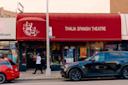 Street view of Thalia Spanish Theatre, with a red awning displaying its name and logo. A person walks by the entrance, and two cars are parked along the curb in front of the building.