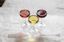 Three coupe glasses filled with red, yellow, and dark red liquids are arranged in a triangle shape on a crumpled white surface.