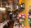 A cozy restaurant with ornate tin ceiling, yellow walls, white tablecloths, wine bottles on shelves, a bouquet of pink roses in a vase, and warm lighting creating an inviting atmosphere.