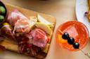 A wooden board with assorted cured meats, sliced pear, and bread, accompanied by a bowl of olives. Next to it is a glass of orange Aperol spritz garnished with black olives on a skewer.