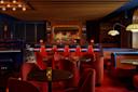 A cozy, dimly lit bar with retro red chairs and round tables. The bar is lined with blue barstools, and a variety of bottles are displayed on shelves. Warm lighting and a wooden ceiling create an inviting atmosphere. A piano is visible on the side.