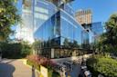 Modern glass building with reflections of the sky and surrounding structures. Patio area with outdoor seating, tables, and chairs partially visible. Lush greenery, bushes, and blooming flowers frame the scene. Bright, sunny day.