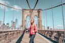 A woman in a red jacket stands on the Brooklyn Bridge with New York City skyscrapers in the background under a clear blue sky.