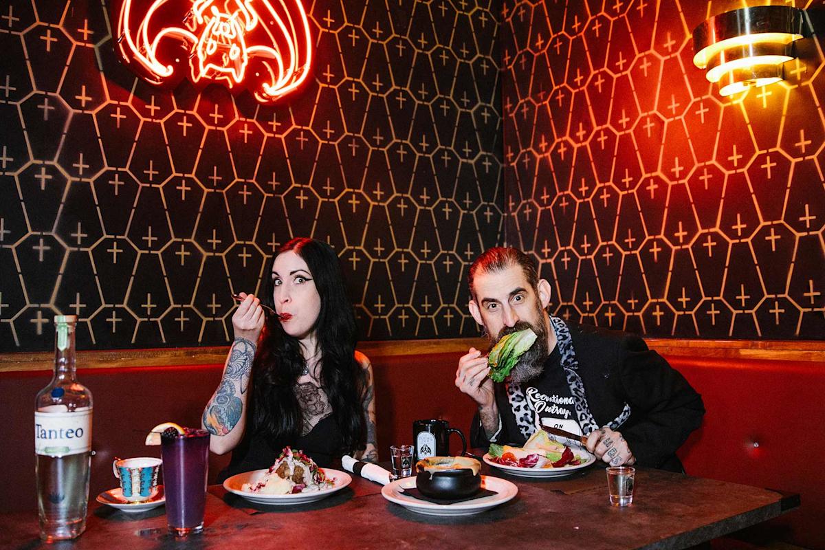 A tattooed pair sits in a dimly lit restaurant booth, eating dinner beneath a red neon bat sign and patterned wall. Drinks and plates of food are on the table.