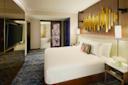 A modern hotel room with a large bed, abstract gold wall art, and a decorative pillow. There's a large TV on one wall, a blue-patterned carpet, a bedside table with a vase of flowers, and an open view into a bathroom.