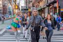 A group of people, including two drag queens in colorful outfits, joyfully walk across a city crosswalk, surrounded by others smiling and laughing on a lively street with rainbow flags and cafes in the background.