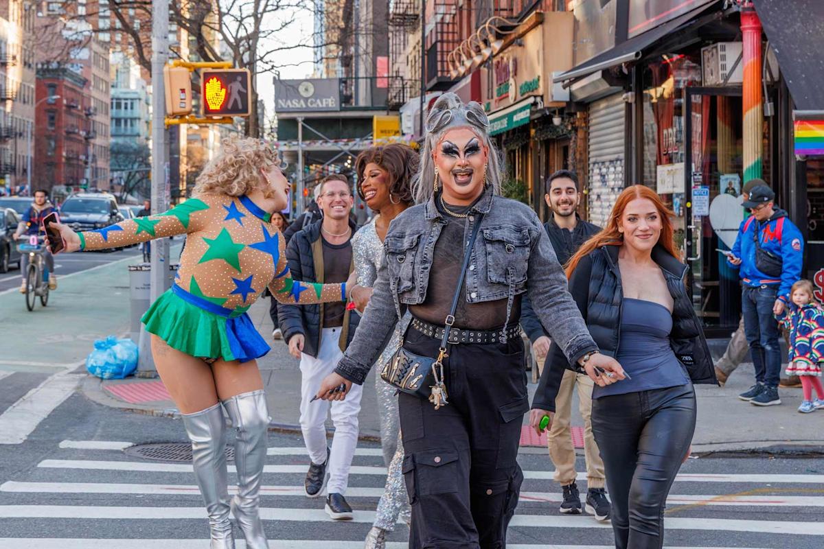 A group of people, including two drag queens in colorful outfits, joyfully walk across a city crosswalk, surrounded by others smiling and laughing on a lively street with rainbow flags and cafes in the background.