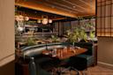 A modern restaurant interior with green leather booths, a wooden table set for four, lush green plants, and warm ambient lighting from round ceiling fixtures. The space has a cozy and inviting atmosphere.