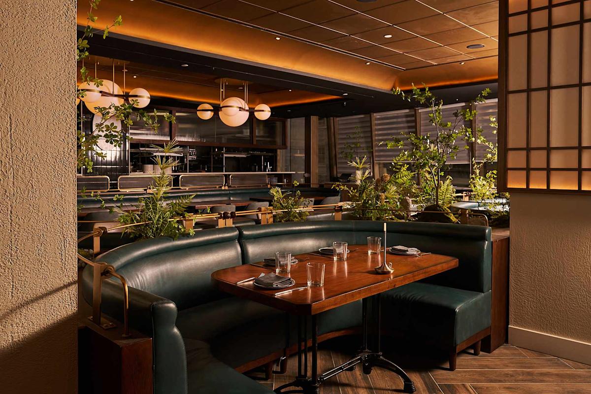 A modern restaurant interior with green leather booths, a wooden table set for four, lush green plants, and warm ambient lighting from round ceiling fixtures. The space has a cozy and inviting atmosphere.