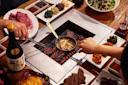 Two people grill meat and mushrooms over a charcoal grill built into a table, surrounded by dishes of raw meat, sauces, kimchi, salads, a bottle of sake, and various side dishes.