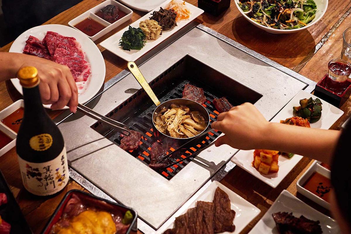 Two people grill meat and mushrooms over a charcoal grill built into a table, surrounded by dishes of raw meat, sauces, kimchi, salads, a bottle of sake, and various side dishes.