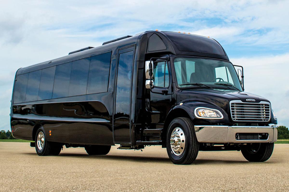 A large black luxury shuttle bus is parked on an open paved area under a partly cloudy sky, with trees and grass visible in the background.