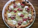 A freshly baked pizza on a white plate, topped with slices of red meat, dollops of white cheese, microgreens, and a drizzle of creamy sauce, sitting on a wooden table.