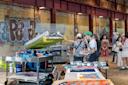 People gather around a screen printing setup in an industrial space with graffiti on the walls. Various colorful prints and equipment are displayed on tables. The atmosphere is lively and artistic.