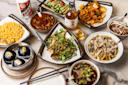 A variety of Asian dishes are artistically arranged on a table. The spread includes savory stir-fries, spicy soups, sesame-topped noodles, marinated meats, and colorful vegetable dishes, with a bottle of liquor and a glass nearby.