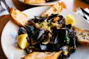 A white plate of steamed mussels garnished with chopped herbs and onions, served with lemon wedges and slices of crusty bread. A fork and knife are visible on the side.