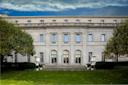 The image shows the facade of The Frick Collection, a grand white neoclassical building with tall windows and ornate details. It is flanked by neatly trimmed bushes and trees under a partly cloudy blue sky.