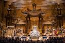 A grand opera stage features ornate, golden, Asian-inspired architecture. Performers in elaborate costumes surround a central figure seated on a throne, while others stand or kneel, creating a dramatic, theatrical scene.