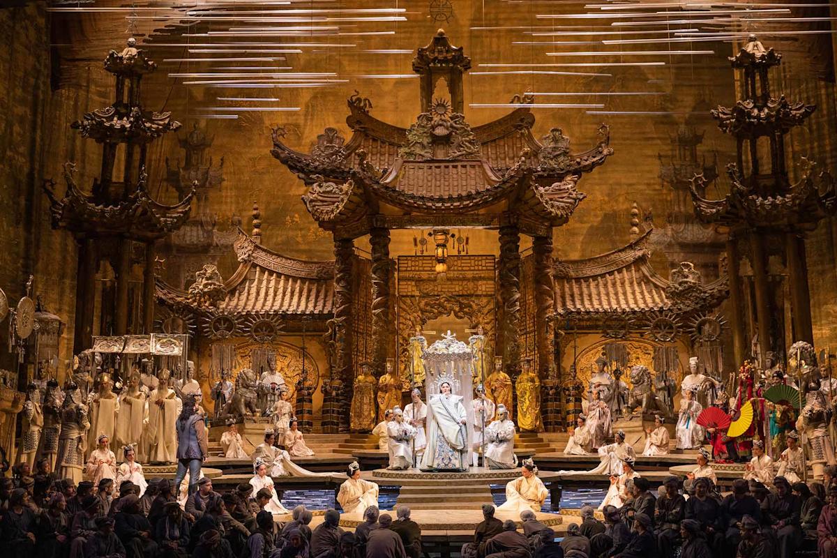 A grand opera stage features ornate, golden, Asian-inspired architecture. Performers in elaborate costumes surround a central figure seated on a throne, while others stand or kneel, creating a dramatic, theatrical scene.