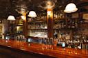 A cozy bar with a polished wooden counter, various liquor bottles and glasses displayed on shelves, warm overhead lights, and vintage-style ceiling tiles, creating an inviting, intimate atmosphere.