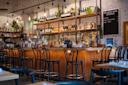 A cozy bar with a wooden counter and high black chairs. Shelves behind the counter display various bottles of wine and spirits. Hanging lights create a warm ambiance. A chalkboard lists drink specials, and potted plants add a touch of greenery.