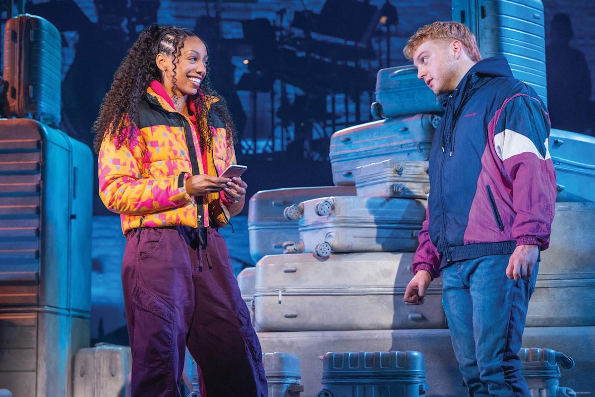 Two actors stand on stage surrounded by suitcases. The woman, smiling, holds a phone and wears a colorful jacket. The man, also smiling, leans slightly toward her, wearing a dark jacket. The background is blue-toned.