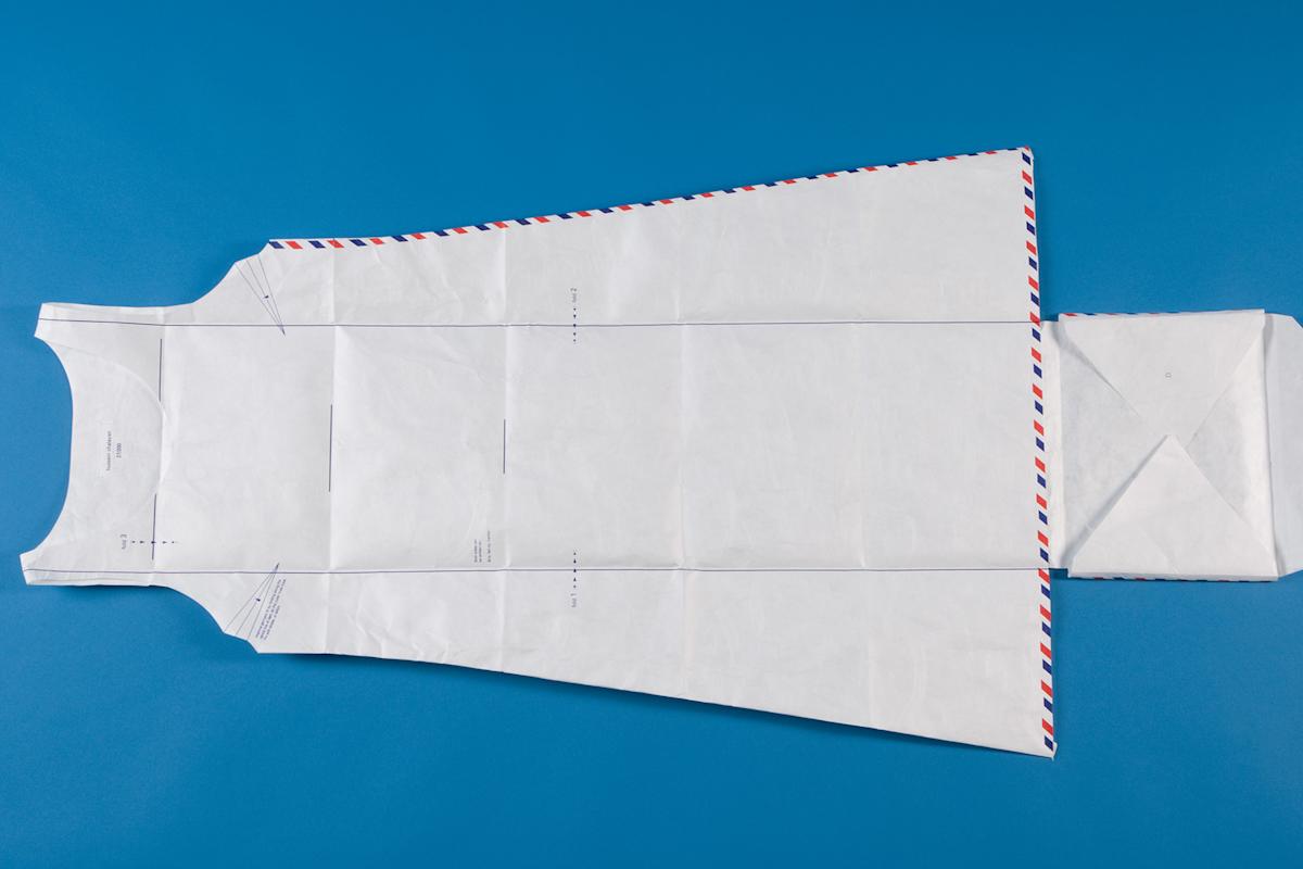 A dress pattern made from white paper with red and blue edging lies flat on a blue background. The pattern includes armholes, neckline, and folded edges, resembling the shape of a sleeveless dress.