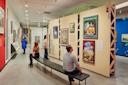 People view vintage travel posters and exhibits in a modern art museum. Two people sit on a bench in the foreground, while others observe displays on the walls. The gallery is brightly lit and spacious.