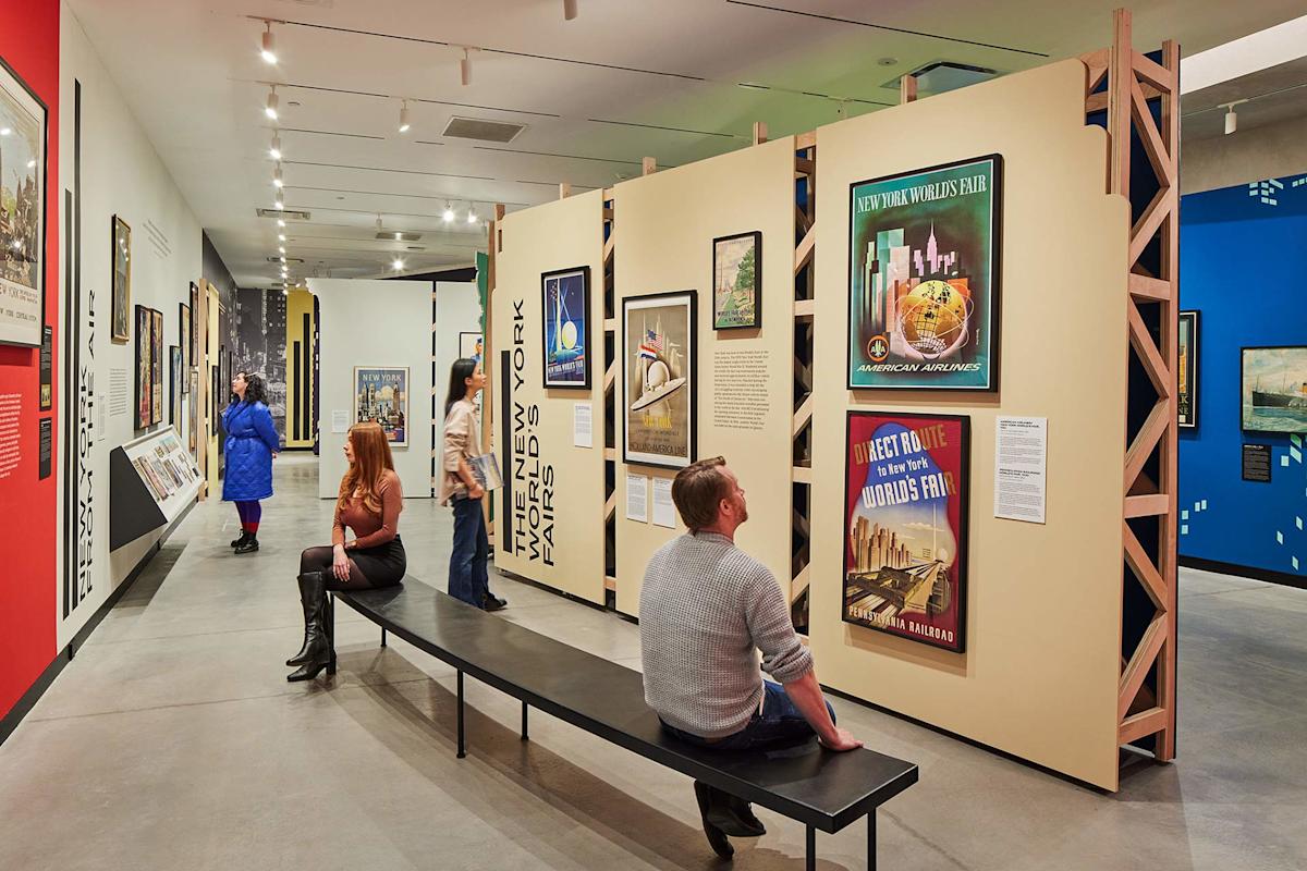 People view vintage travel posters and exhibits in a modern art museum. Two people sit on a bench in the foreground, while others observe displays on the walls. The gallery is brightly lit and spacious.