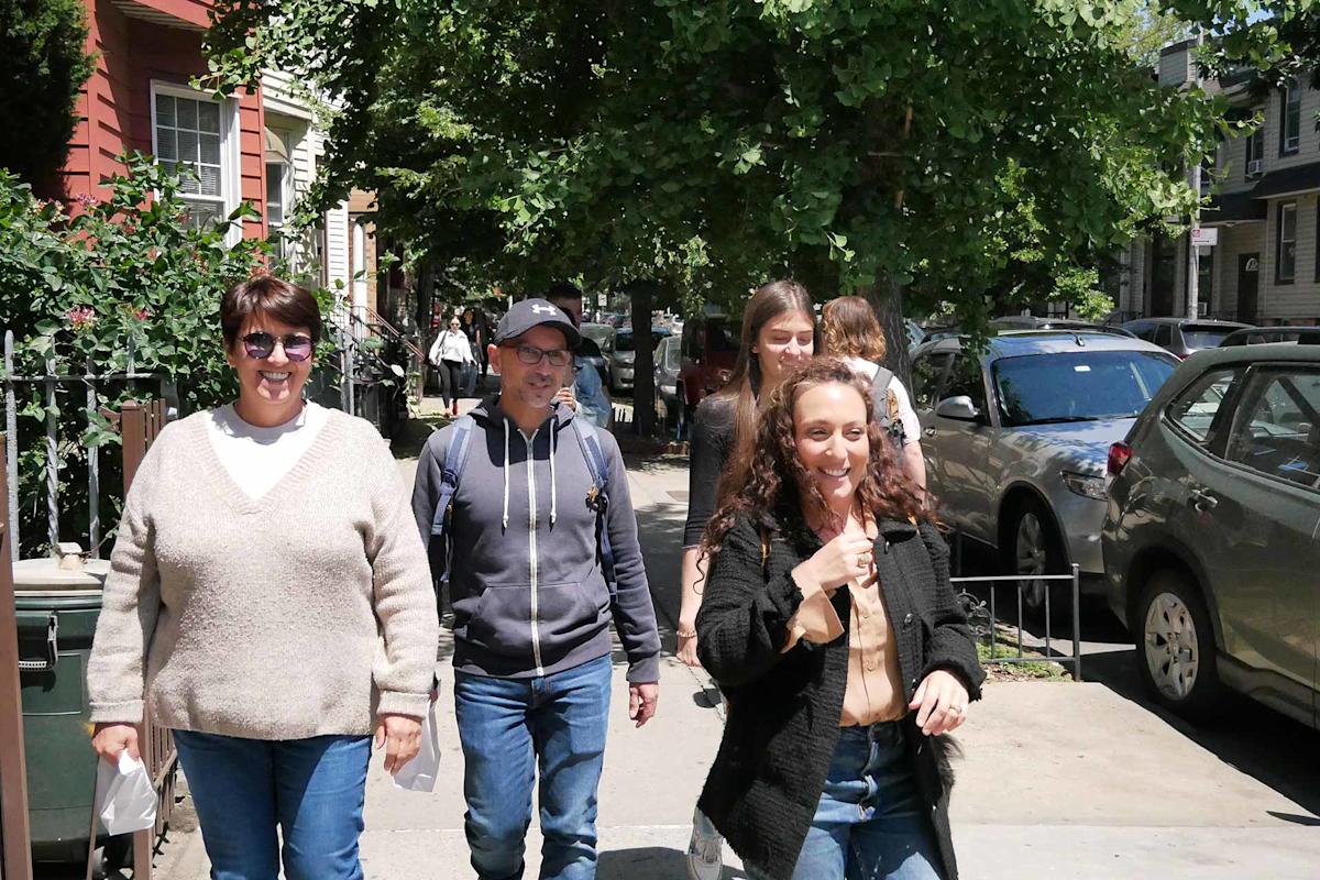 Four people are walking on a sunny sidewalk lined with parked cars and trees, smiling and enjoying the day. Two women lead in front, a man and another woman follow behind. Houses and greenery are visible in the background.