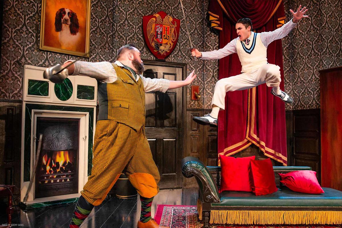 Two actors on a colorful stage set: one in a yellow plaid suit holds a teapot, while another in white leaps mid-air toward him. The background features a fireplace, a green couch, and ornate wallpaper.